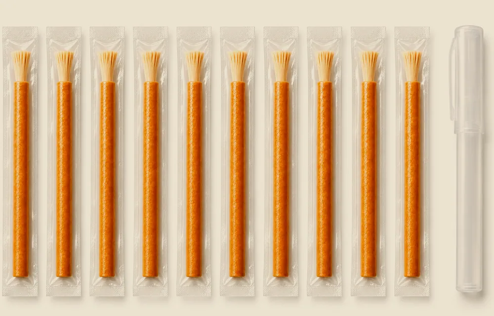 An open carton holding ten individually sealed miswak sticks upright, with further cartons stacked behind and loose sealed sticks alongside
